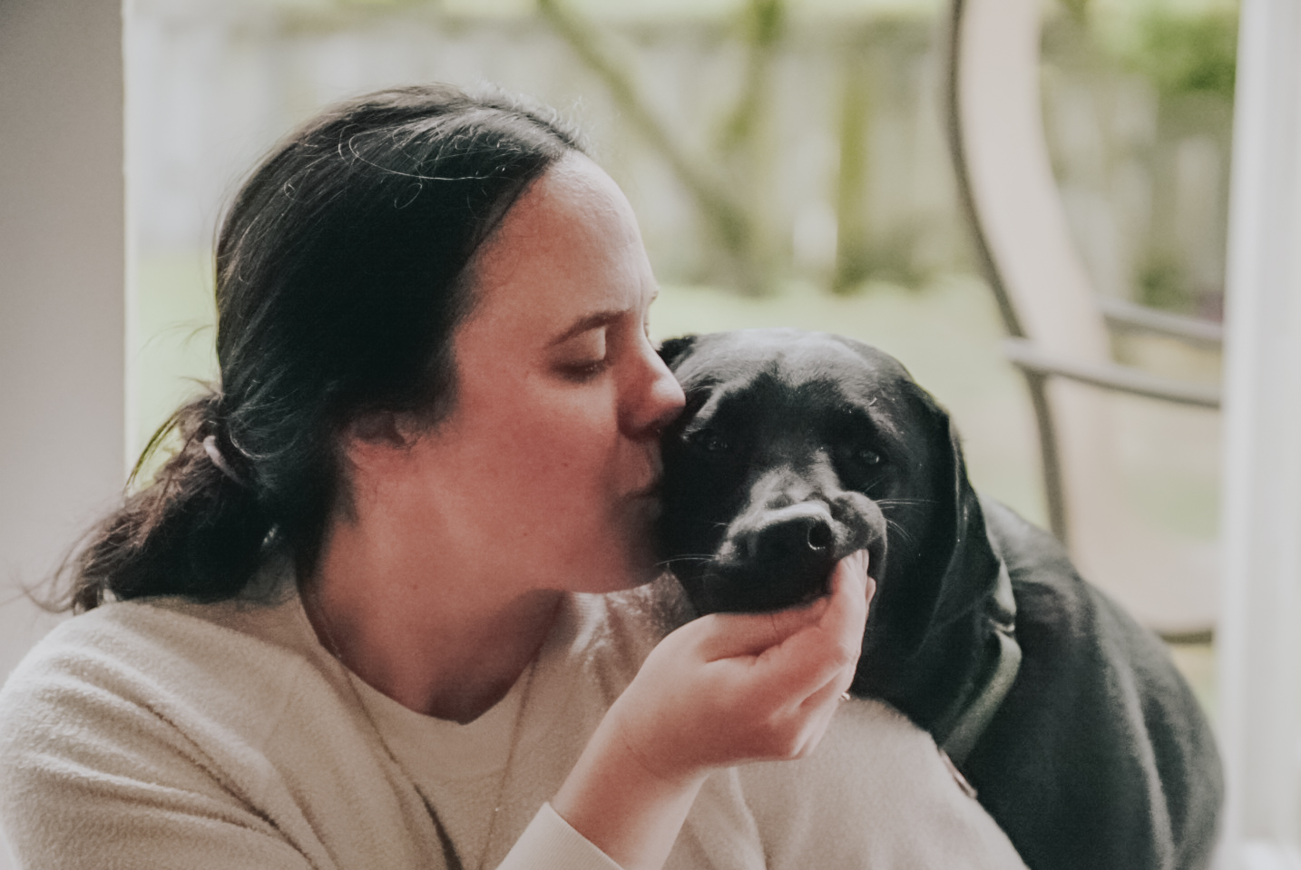 Image of a woman kissing a black lab who is taking a treat from her hand